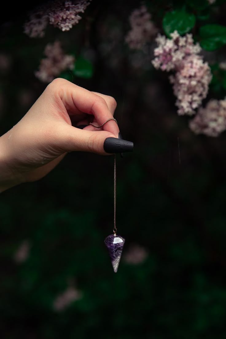 Pendule en améthyste tenu à la main devant un feuillage sombre et des lilas